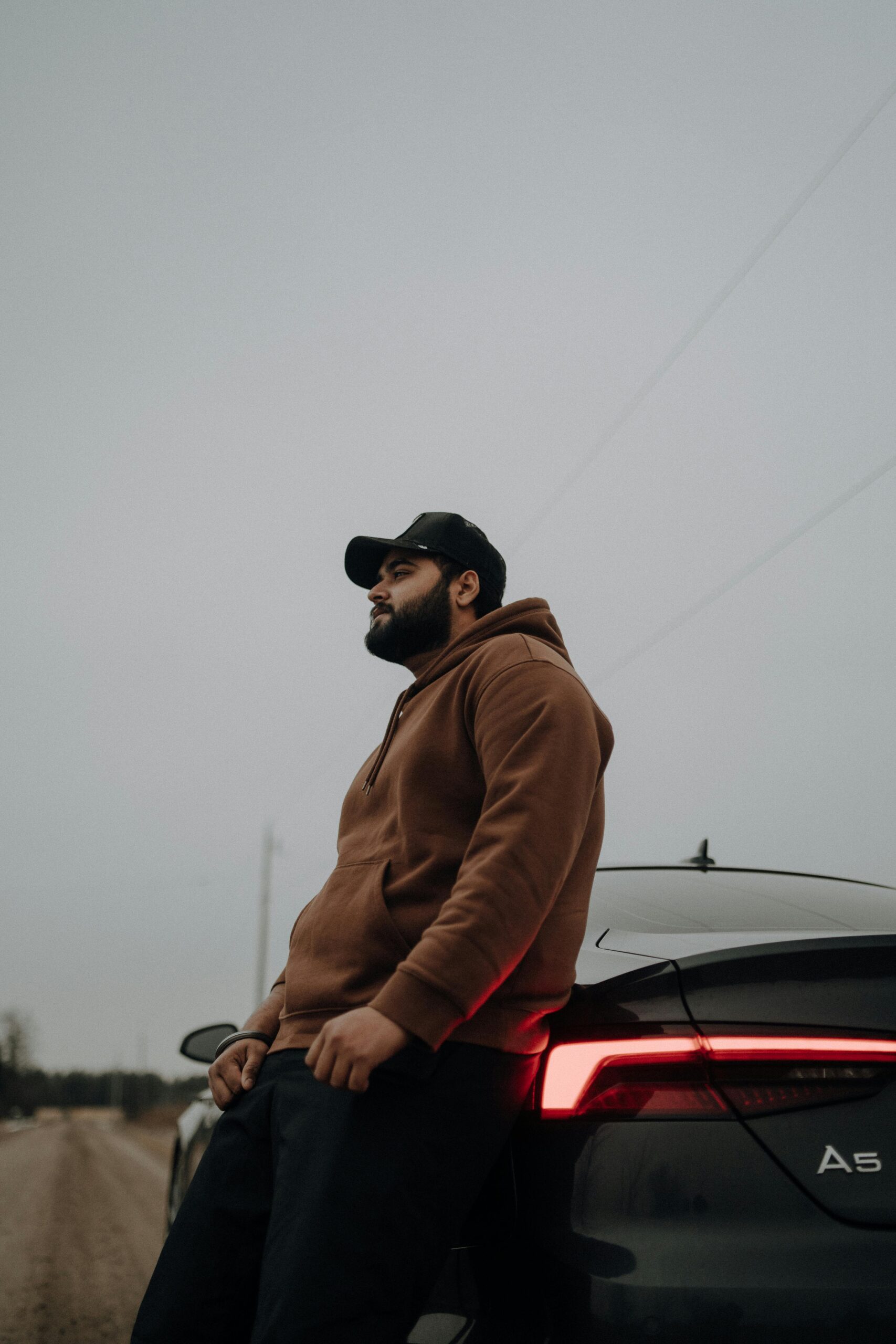 Stylish man in hoodie and cap standing by an Audi A5 on a rural road.